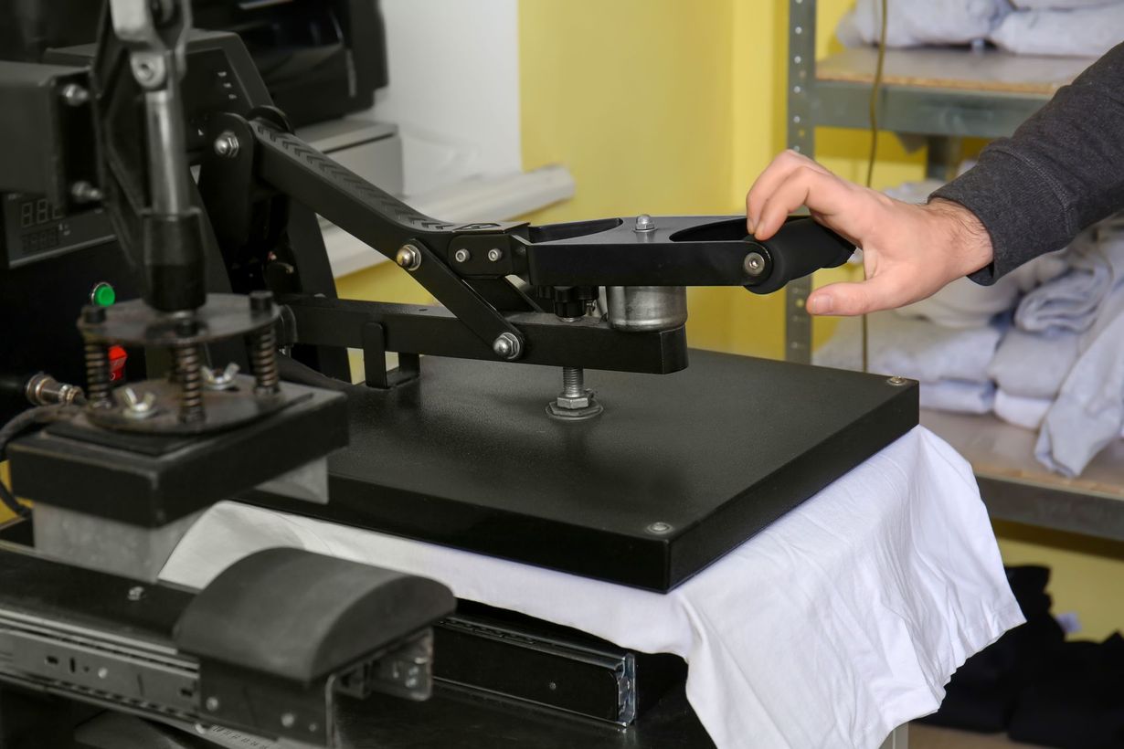 Hand pressing a black heat press machine over a white shirt on a worktable
