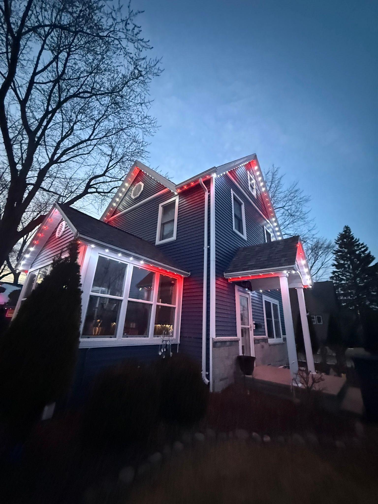 Two-story house at dusk with red holiday lights and a bare tree against a blue sky