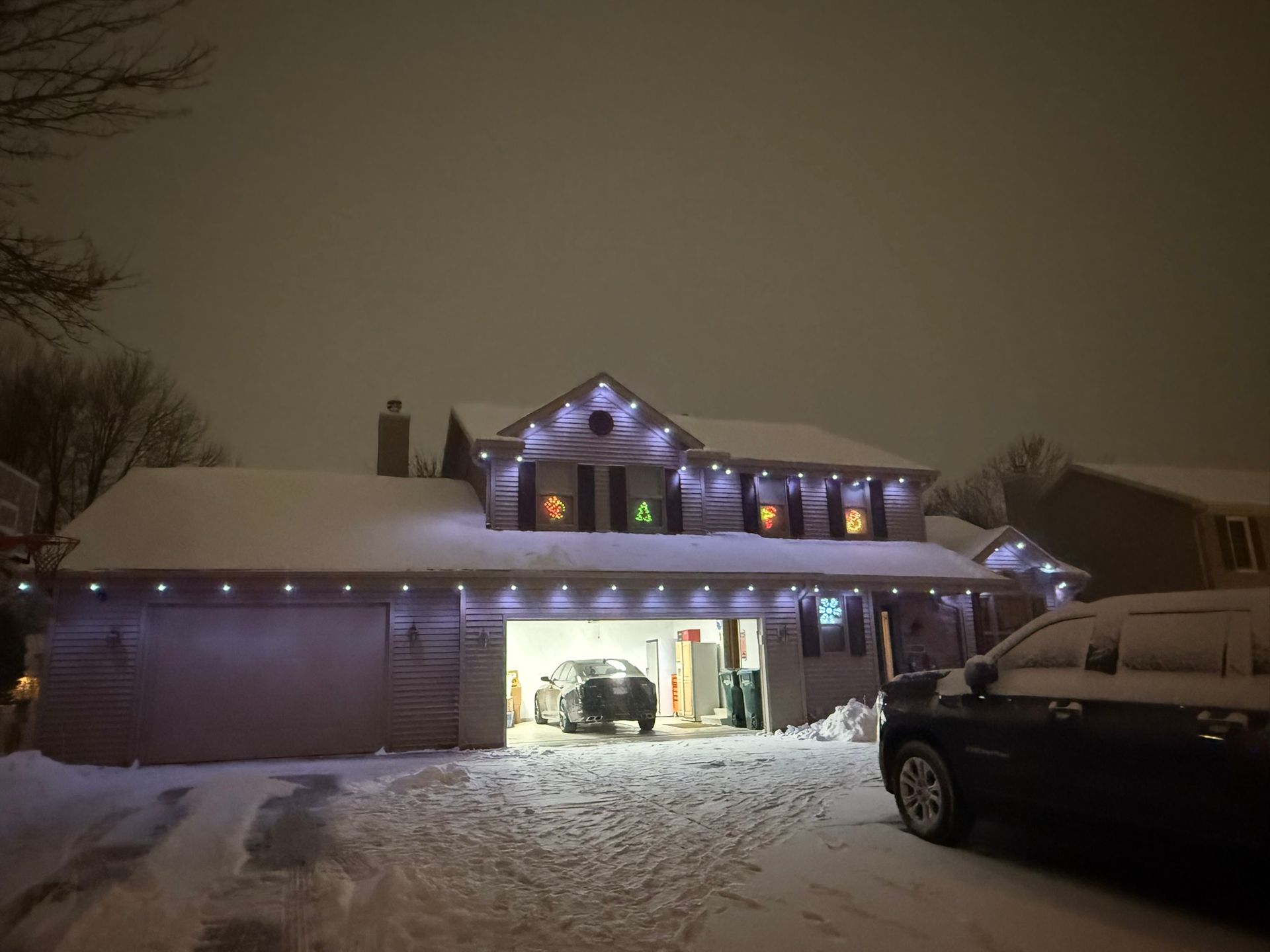 Snowy suburban house at night, lit with purple holiday lights and a car parked in the driveway.
