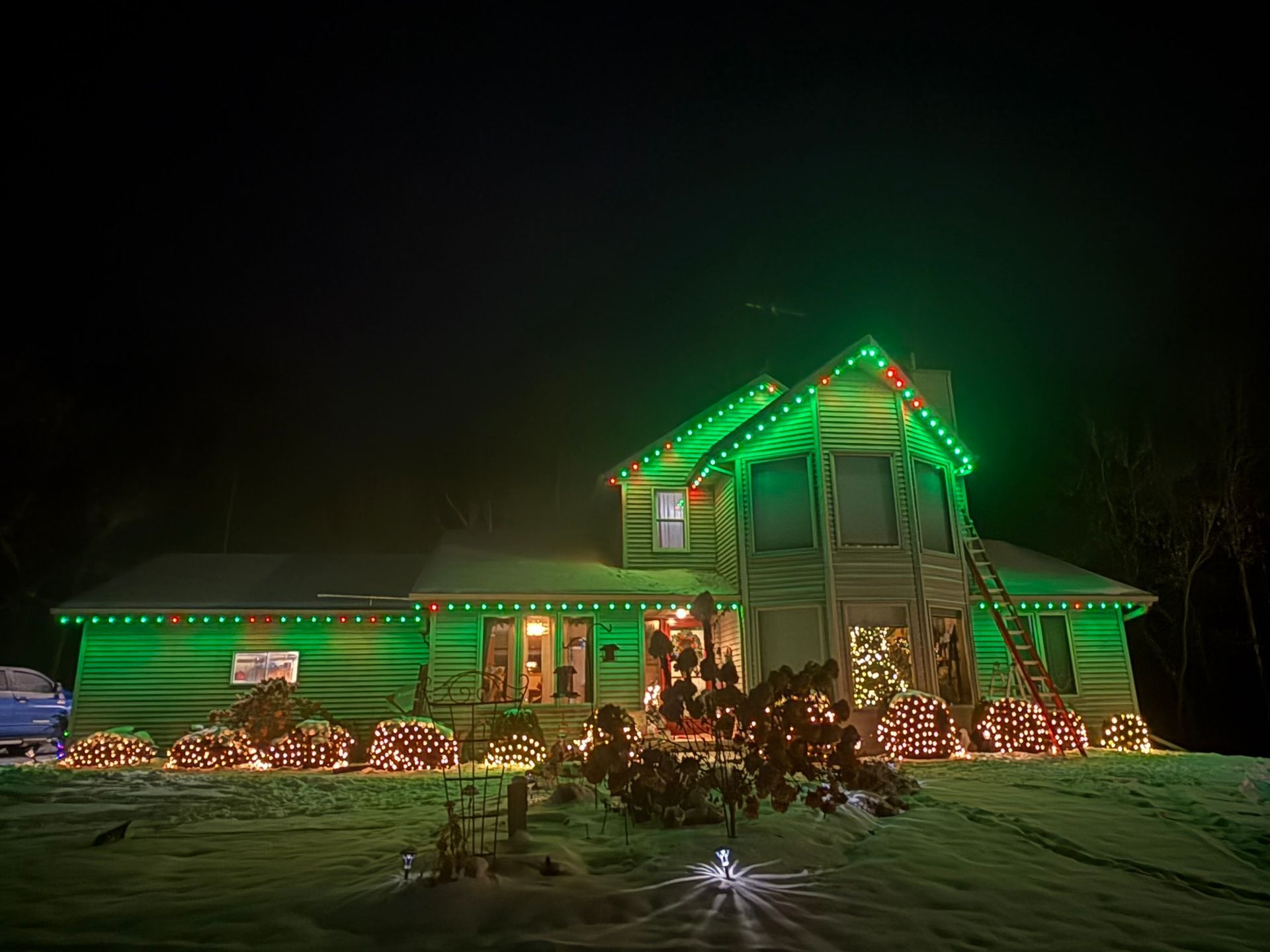 House outlined in green and red Christmas lights at night with snow on the ground