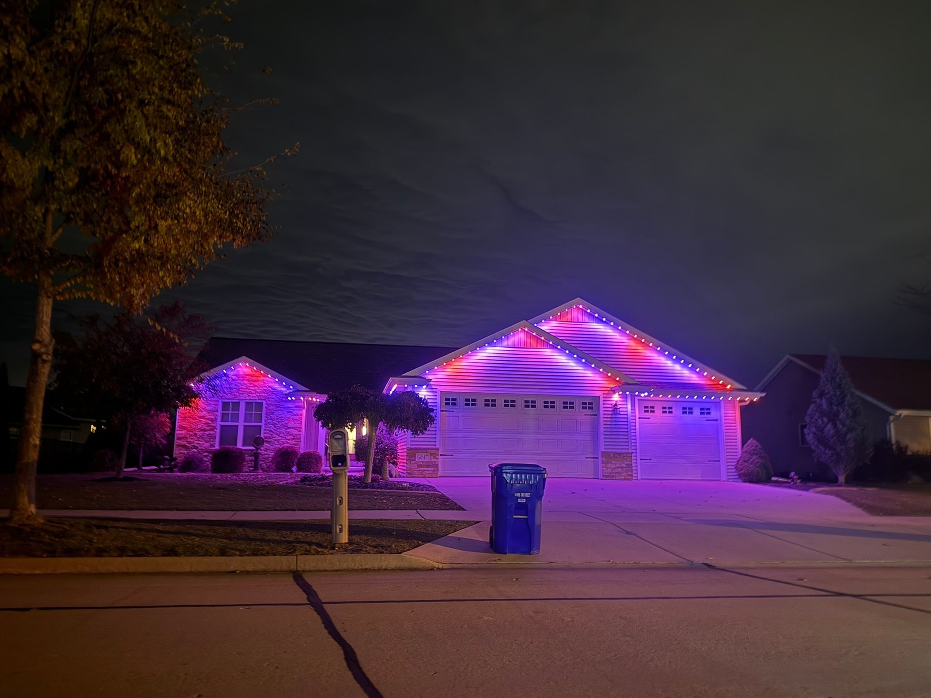 House decorated with purple and pink holiday lights at night, with a blue trash bin in front.