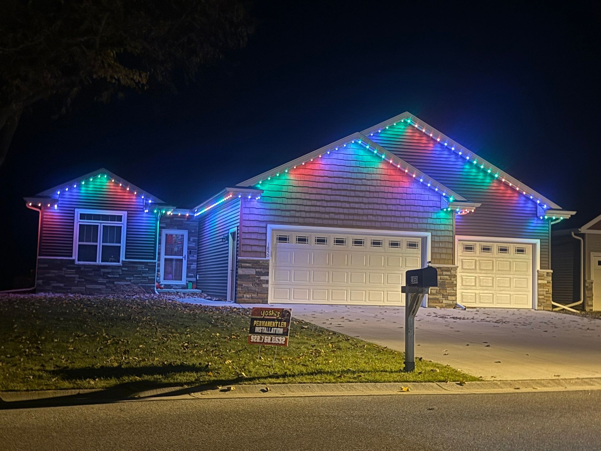 House with colorful Christmas lights along the roofline at night, with two garage doors and a mailbox in front.