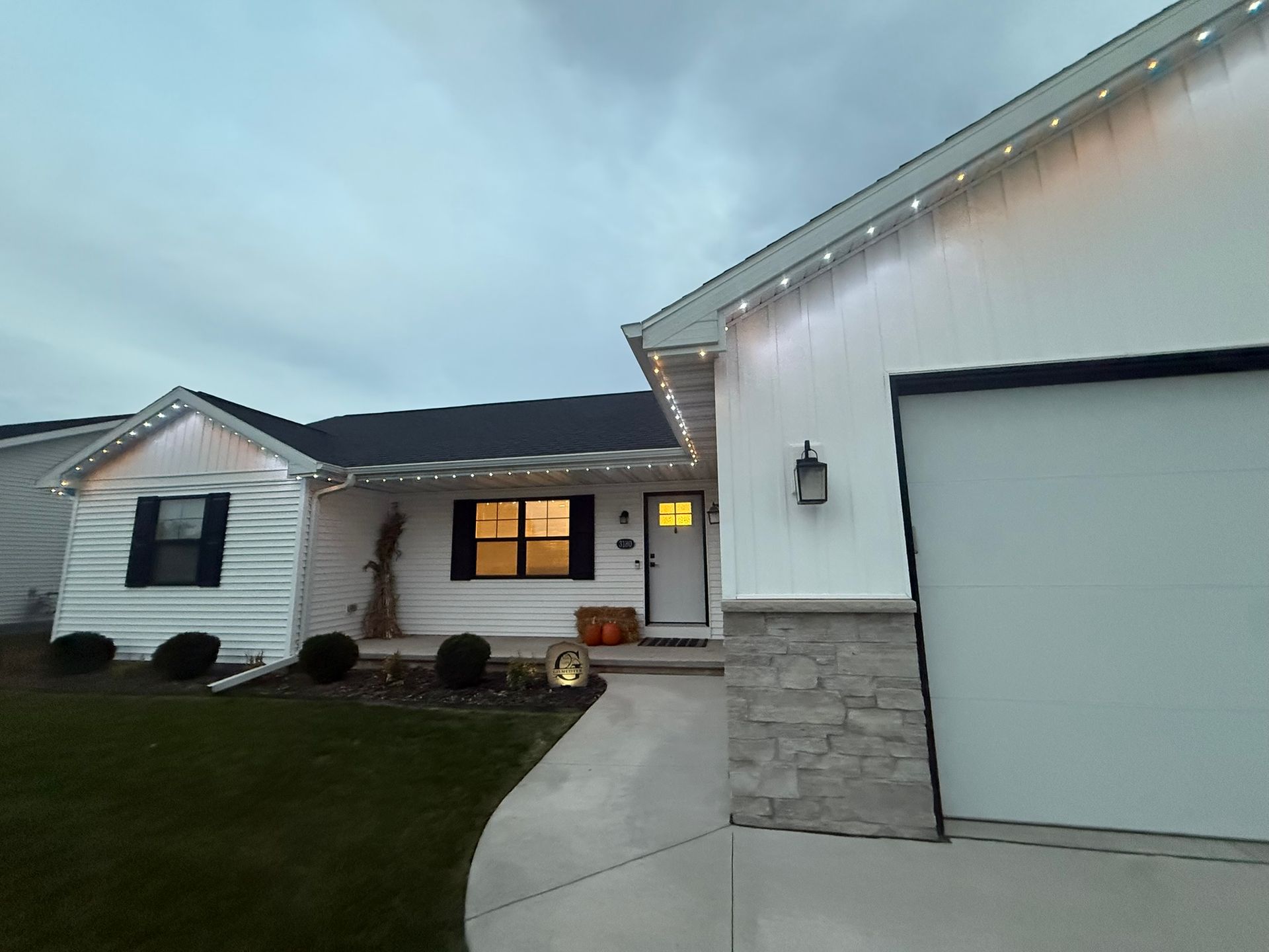 Suburban house exterior at dusk with lit windows, white garage, and a curved concrete driveway.