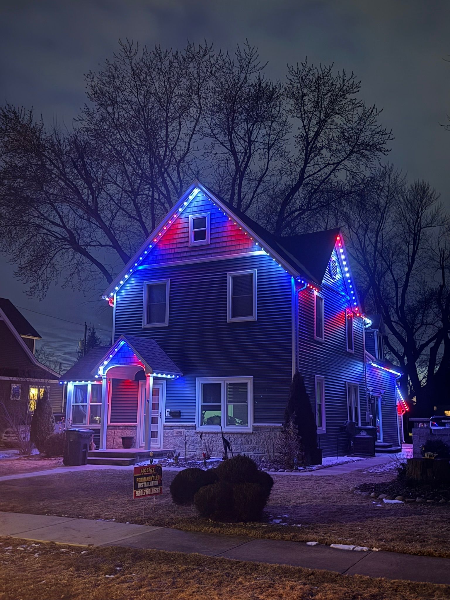 Blue house outlined in red and blue holiday lights at night, with snow on the ground and bare trees behind it
