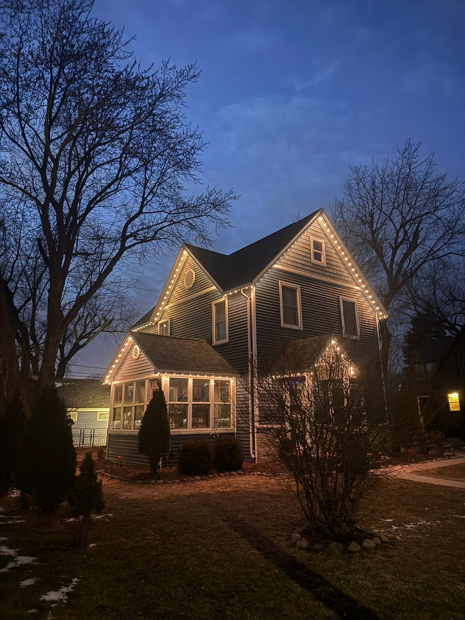 House outlined with warm white holiday lights at dusk, framed by bare trees and a dark blue sky.