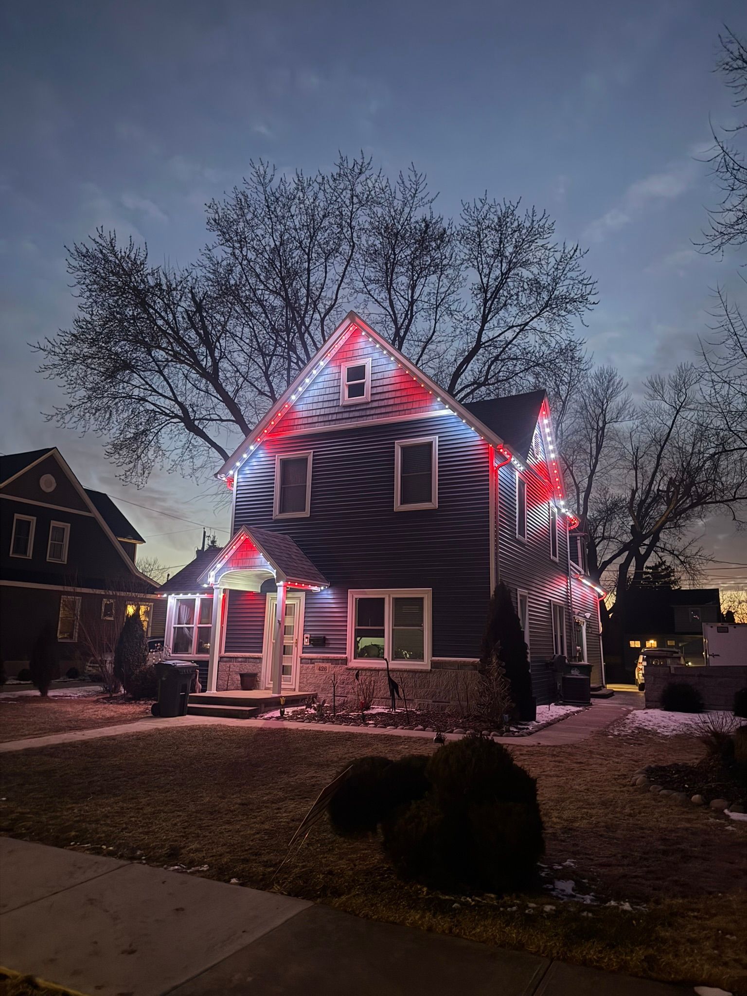 House outlined in red and white holiday lights at dusk, with bare trees and a neighbor’s home nearby