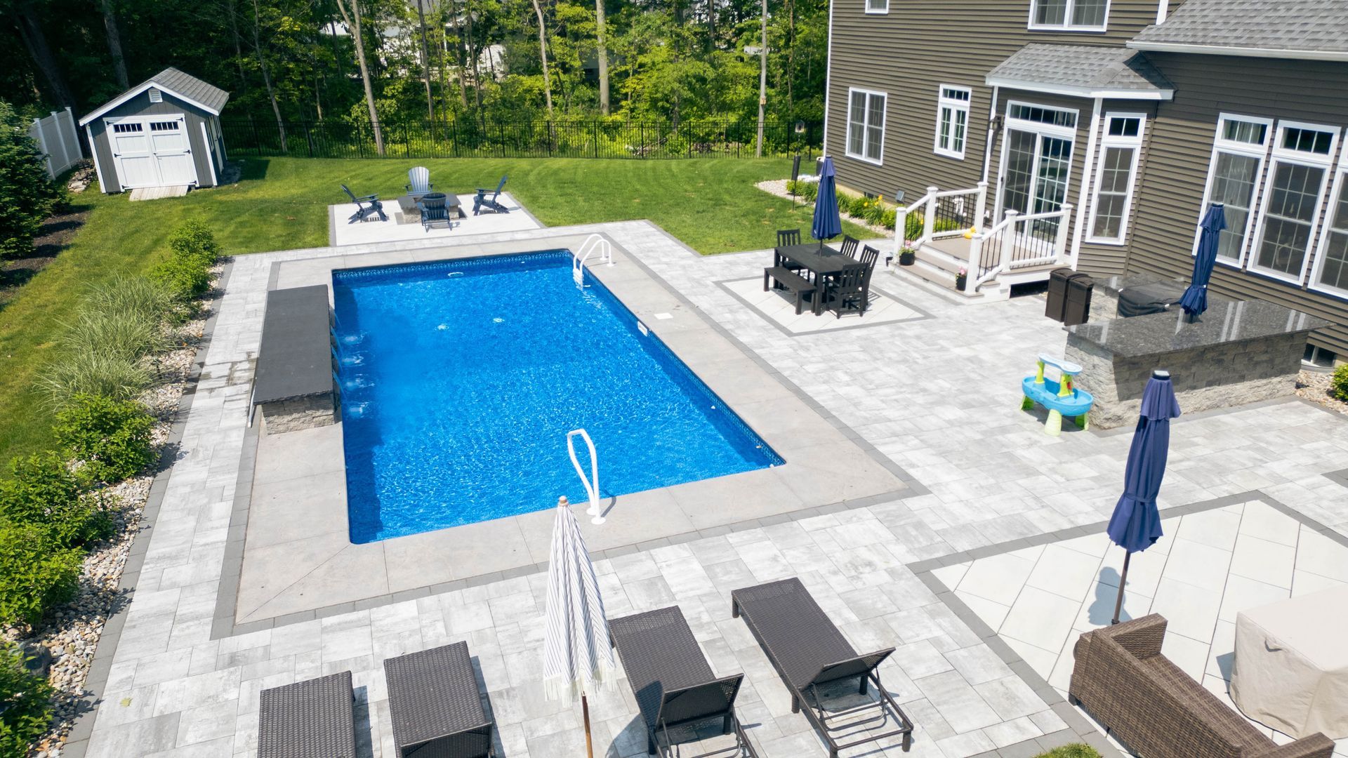 Aerial view of a rectangular pool with patio furniture and a house. Blue water, gray patio, and green lawn.