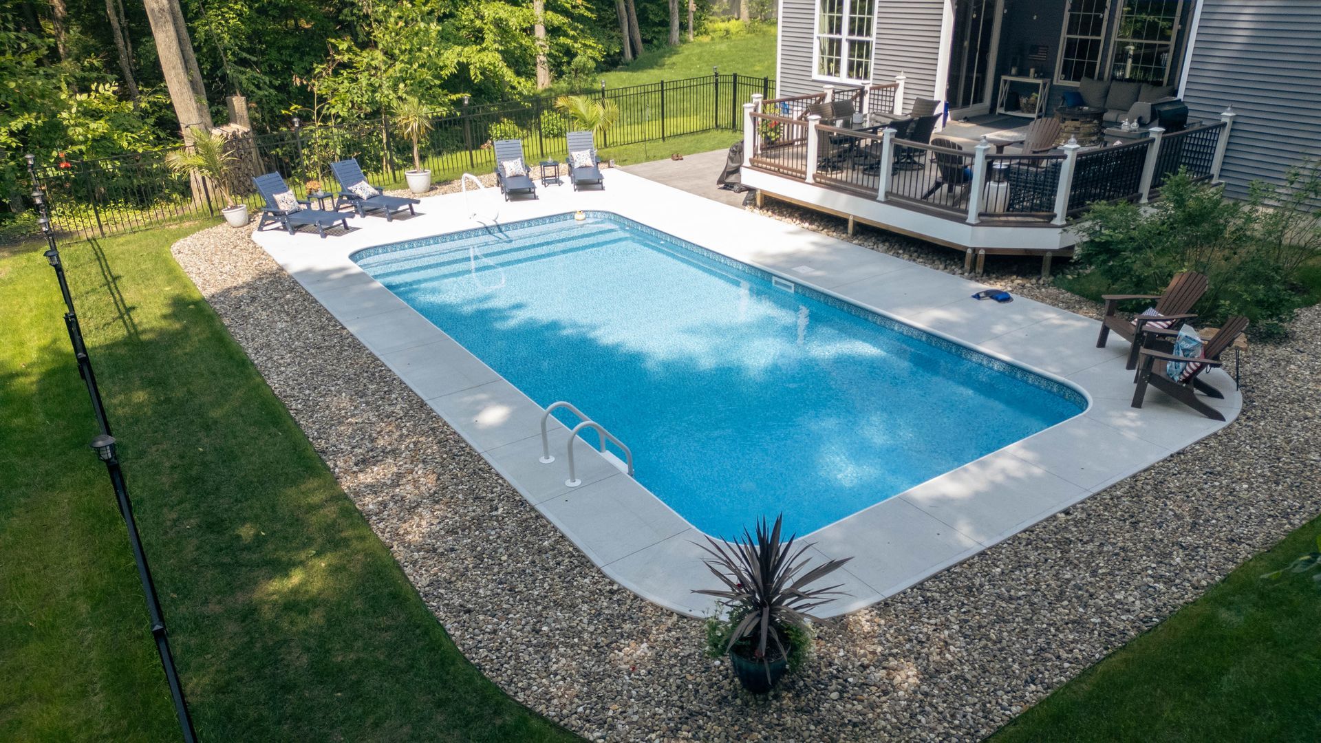 Rectangular in-ground pool surrounded by concrete and gravel, next to a deck and lawn. Chairs sit around the pool.