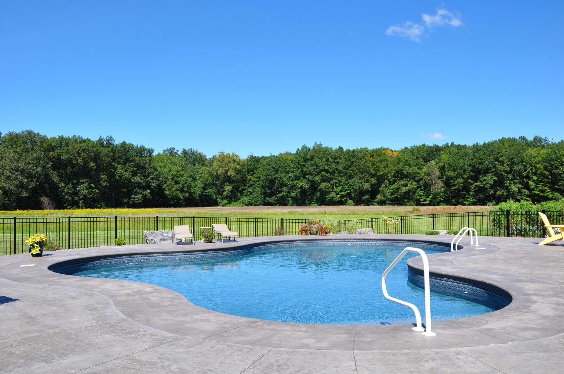 Swimming pool with blue water and surrounding concrete patio, trees in background under a blue sky.