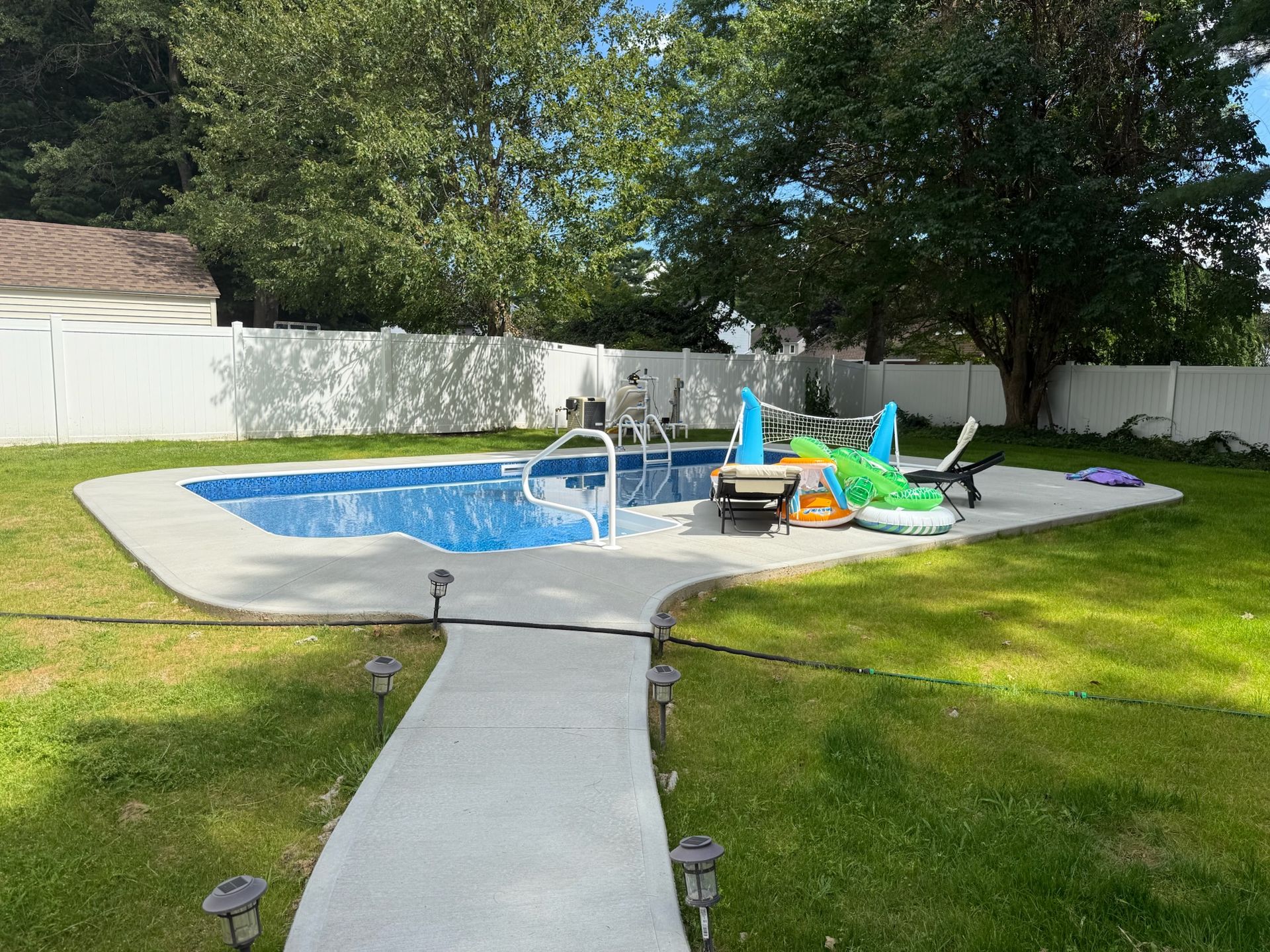 Backyard pool with concrete patio, lounge chairs, and inflatable toys, surrounded by green grass and white fence.