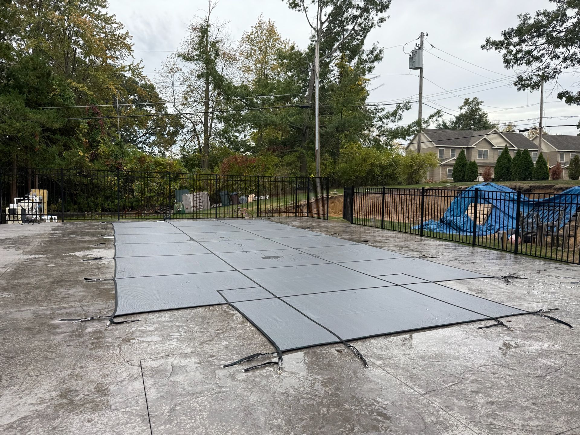 Pool covered with a gray safety cover on a concrete patio, surrounded by a fence and trees.