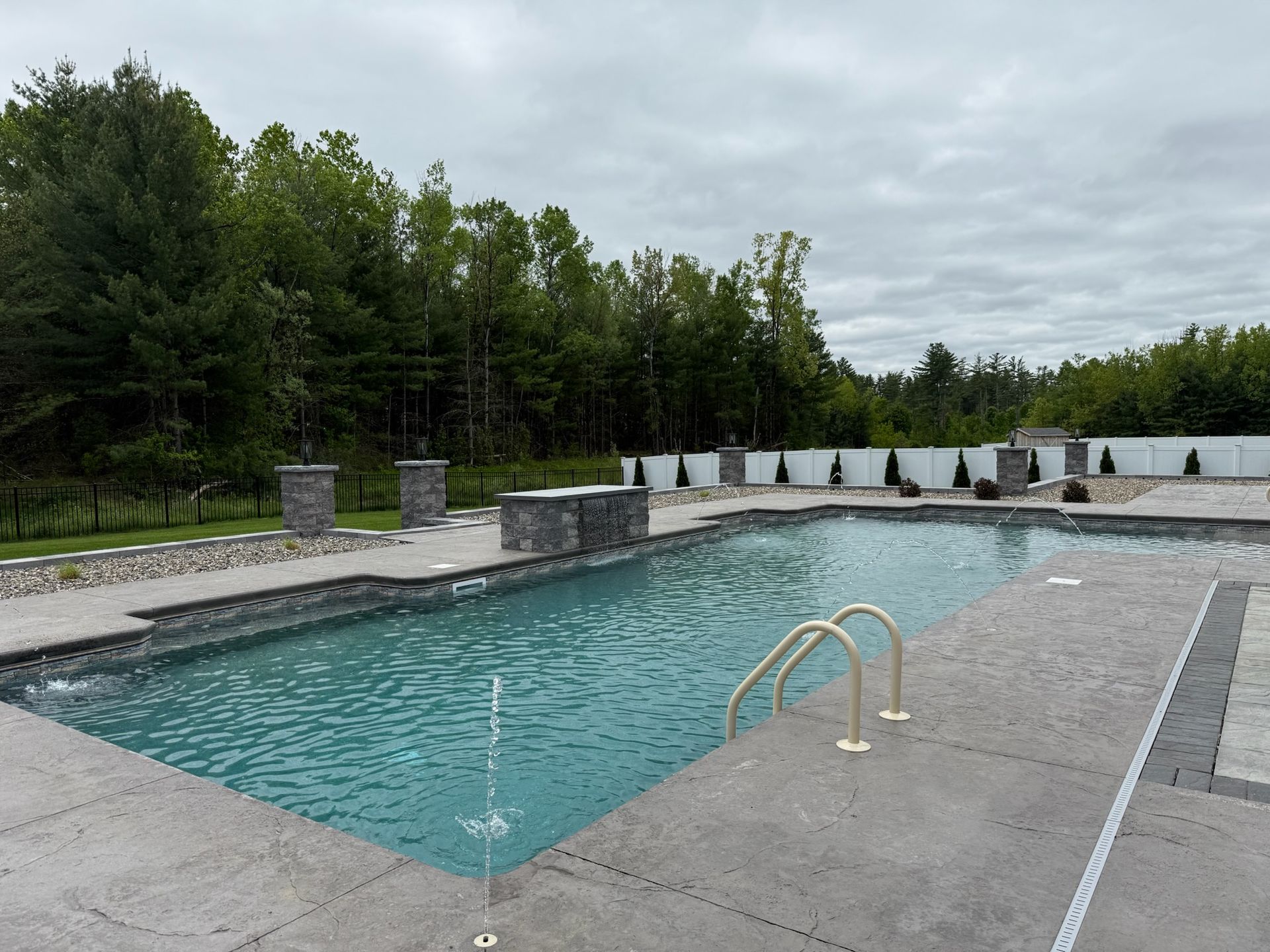 Swimming pool with stone coping, stone accents, and forested background. Cloudy sky.