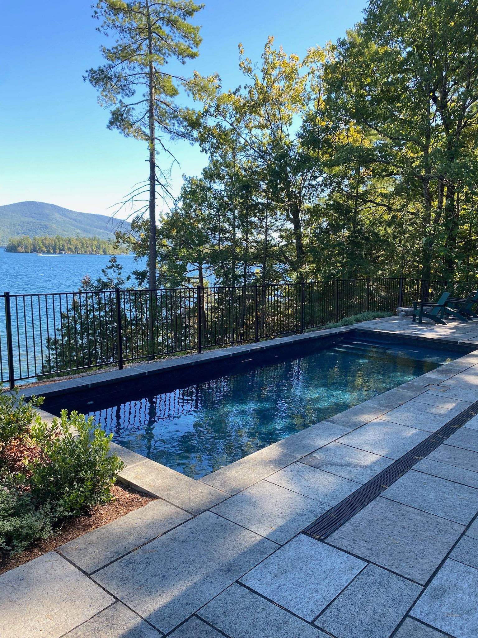 A rectangular pool overlooks a lake with mountains. Blue water, stone patio, black fence, and trees.