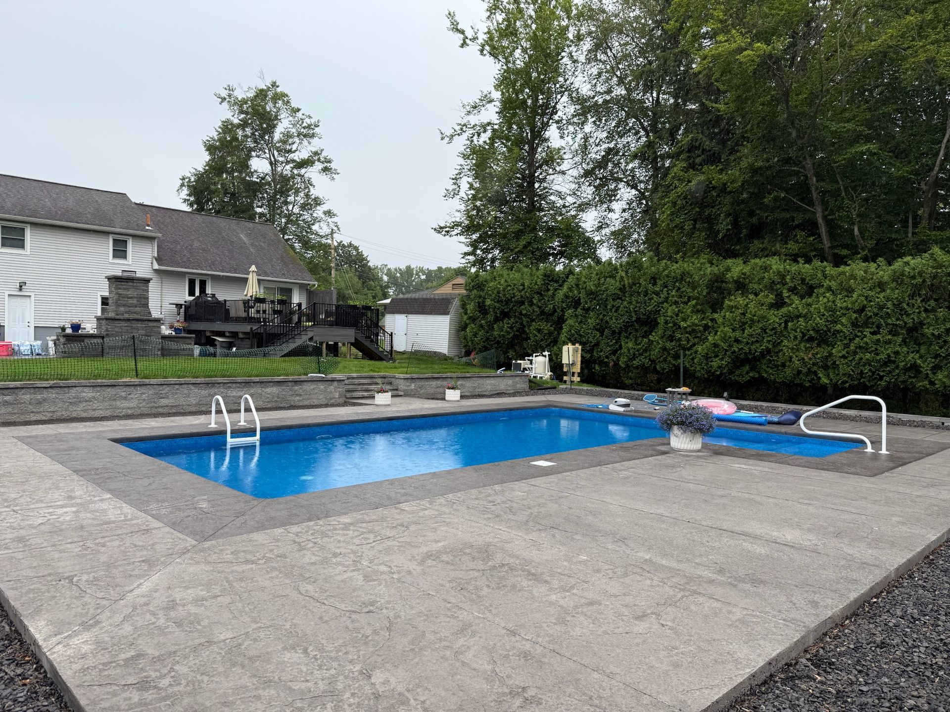 Backyard with a blue rectangular pool surrounded by a concrete patio. A house and trees are in the background.