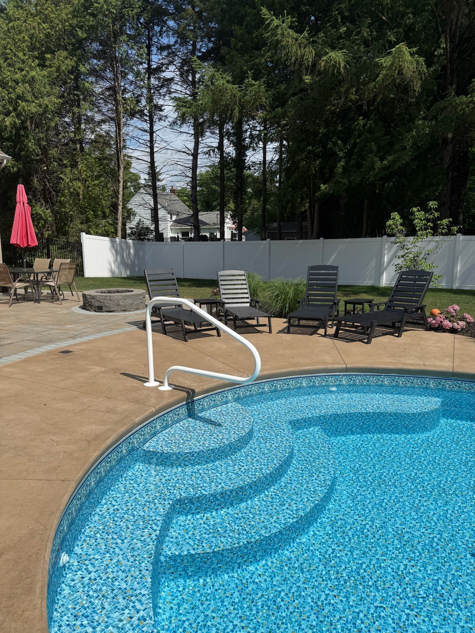 Pool with built-in steps, handrail, and lounge chairs in a backyard setting; trees in the background.