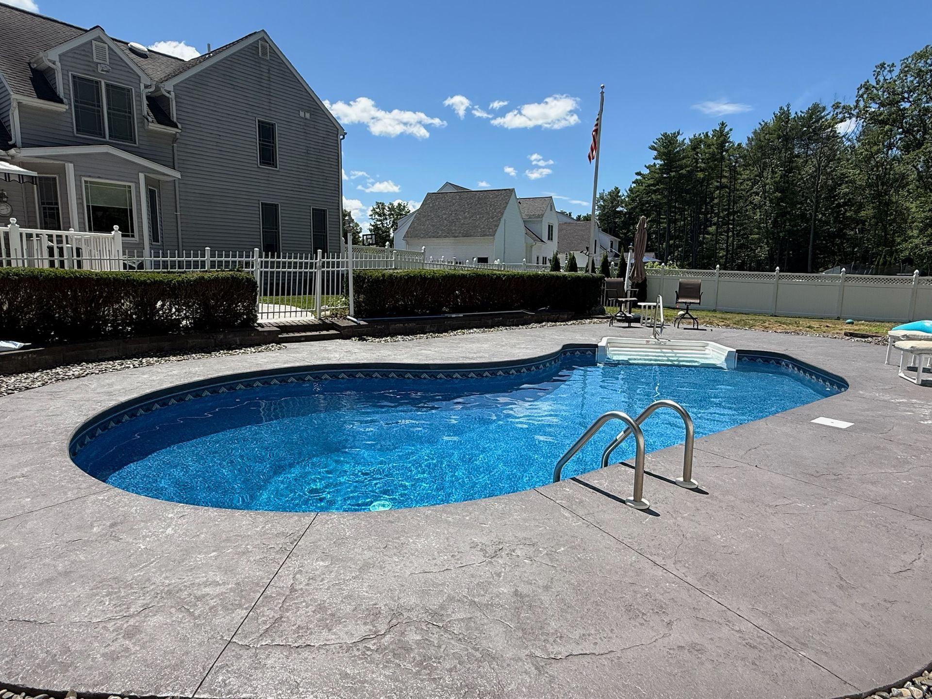 Swimming pool with blue water surrounded by a concrete patio. A two-story house is in the background.
