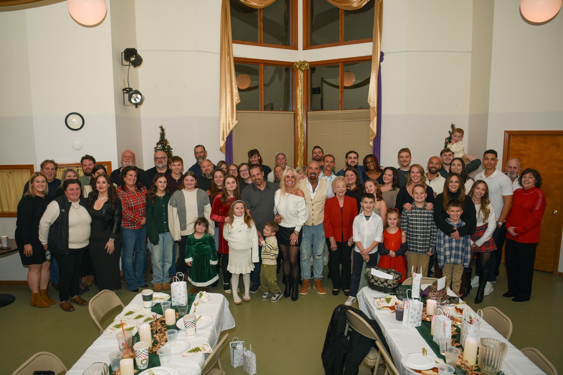 Large group of people gathered at a celebration, tables set, neutral-toned room.