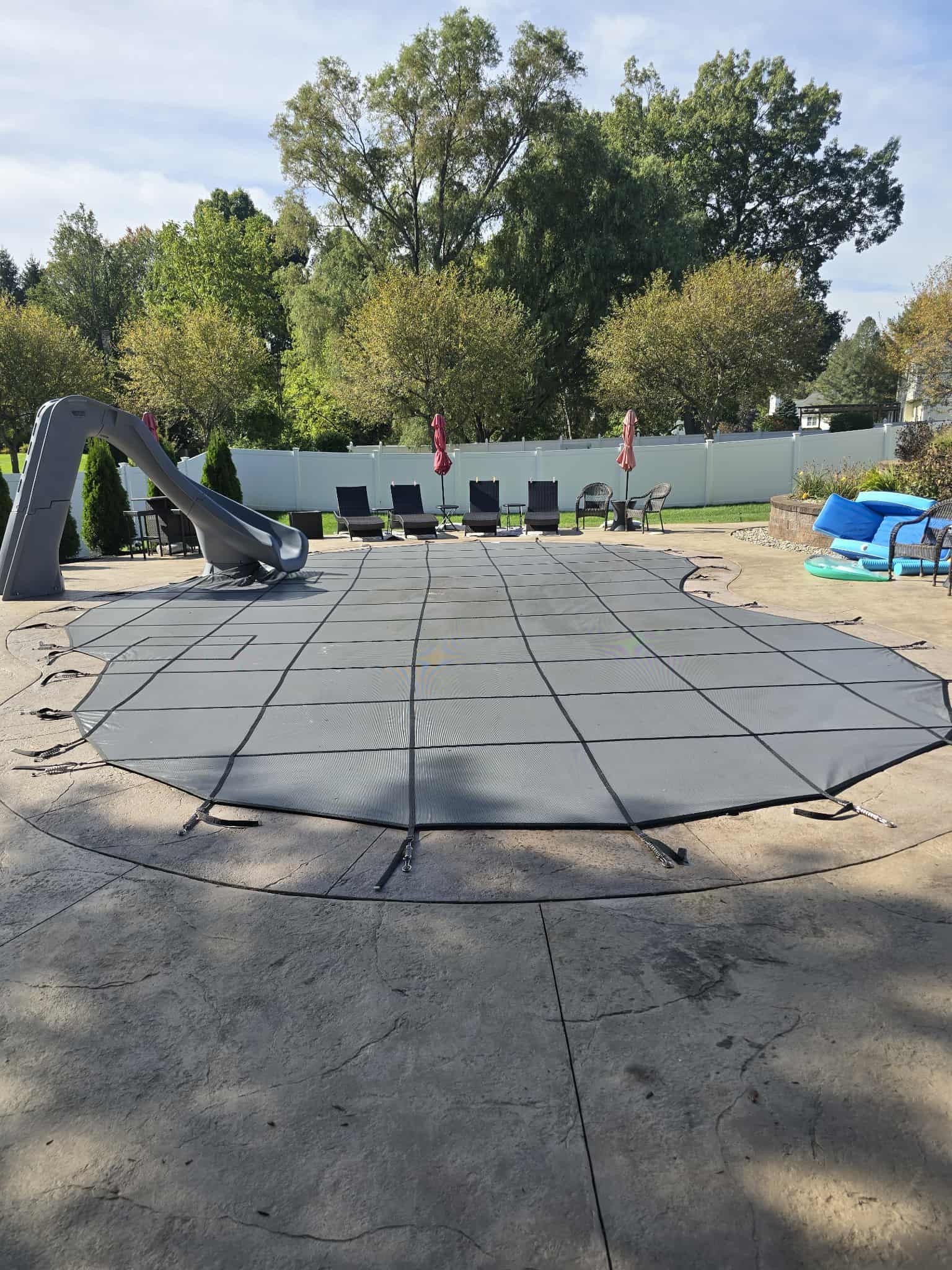 Gray pool cover on concrete patio, slide and lounge chairs in the background. Green trees and blue sky.