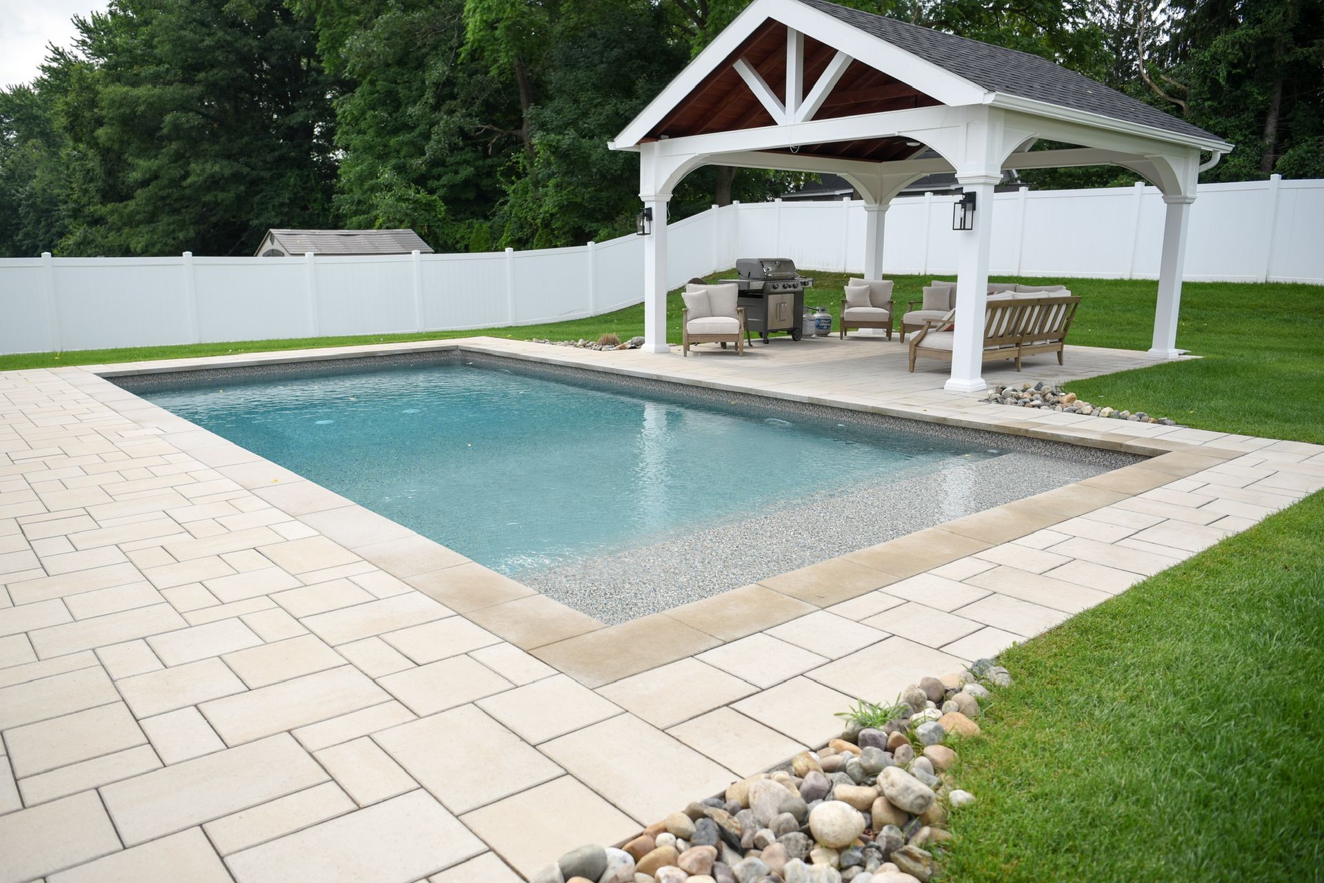 Rectangular pool with a white gazebo, patio furniture, and paver surround in a backyard.