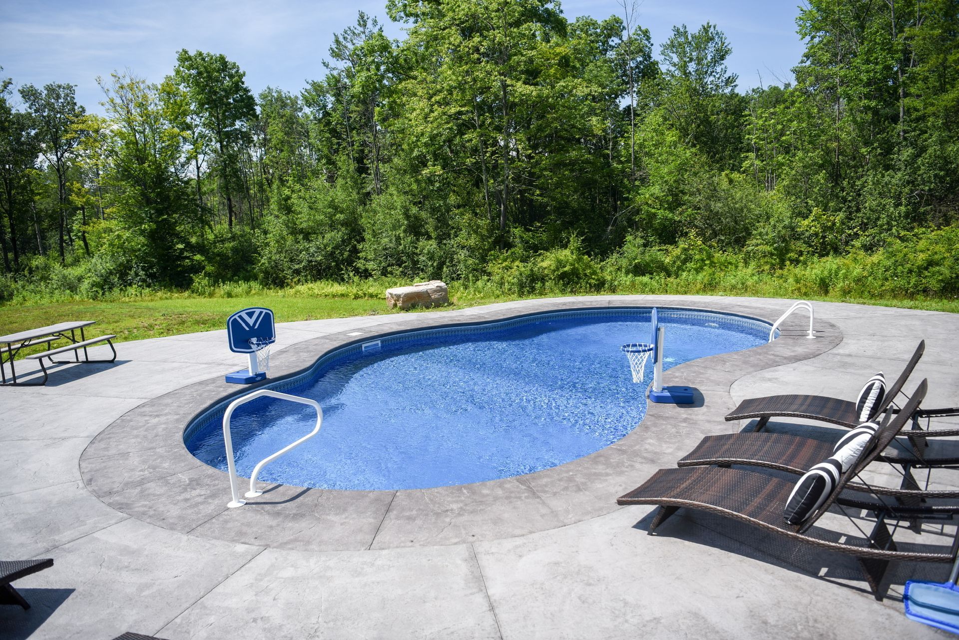 Pool with blue water and concrete patio, surrounded by trees and lawn. Lounge chairs and a picnic table are nearby.