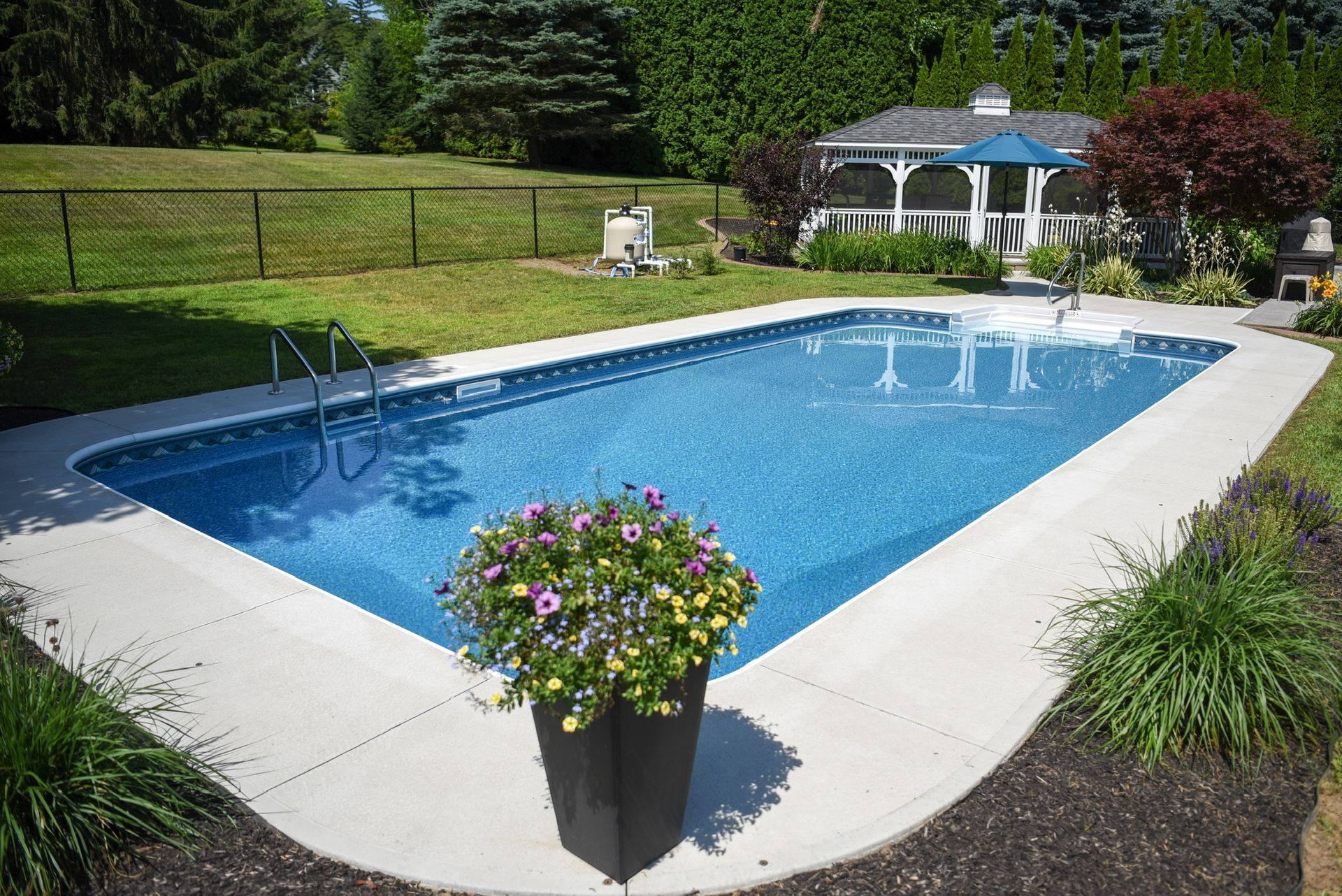 Rectangular pool in a backyard with a gazebo, lush grass, and a planter with colorful flowers.