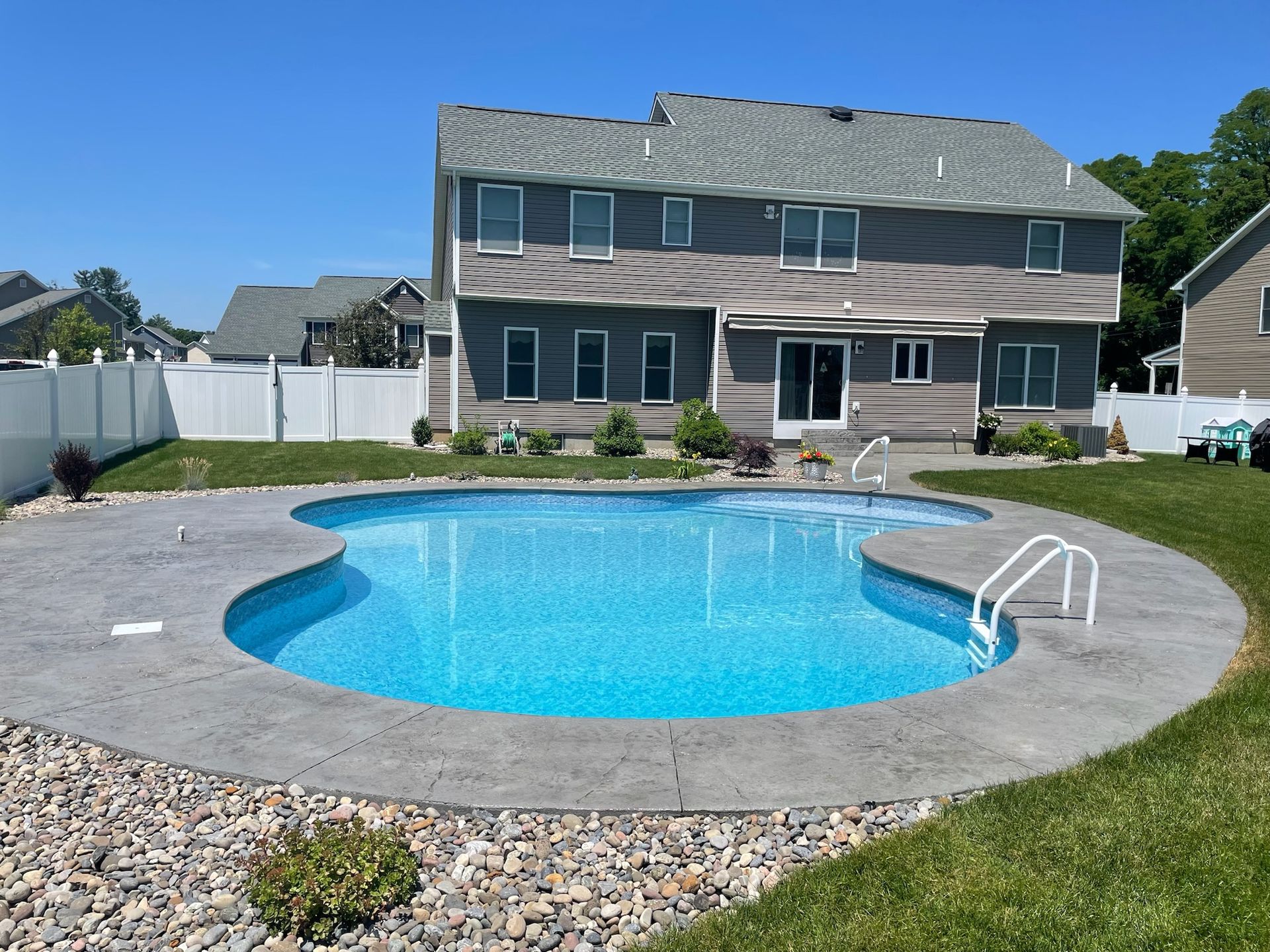 Backyard with a pool in front of a two-story gray house. White fence on left, green grass surrounds pool.