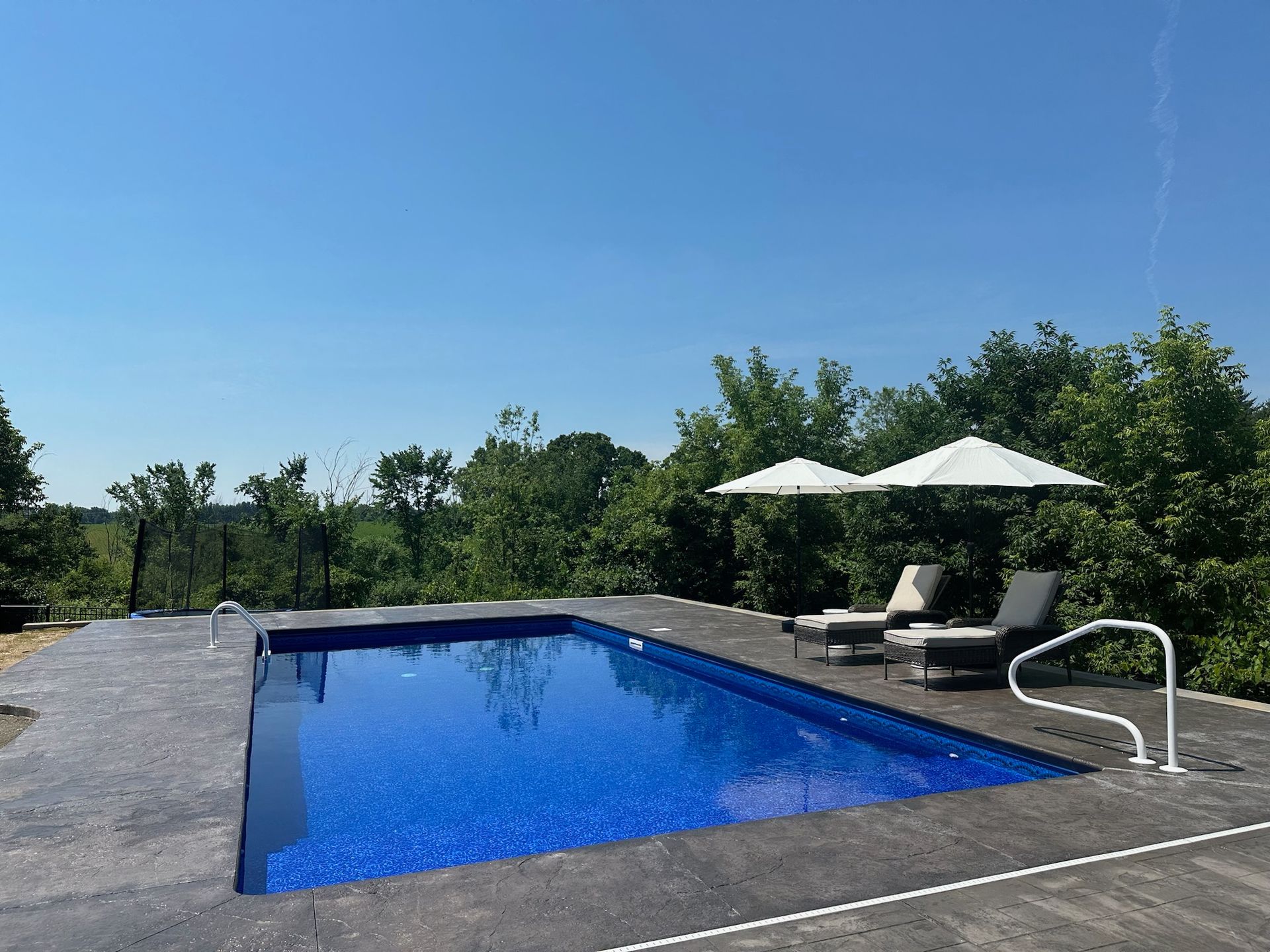 Rectangular pool with blue tile surrounded by gray concrete, two lounge chairs, and two umbrellas on a sunny day.