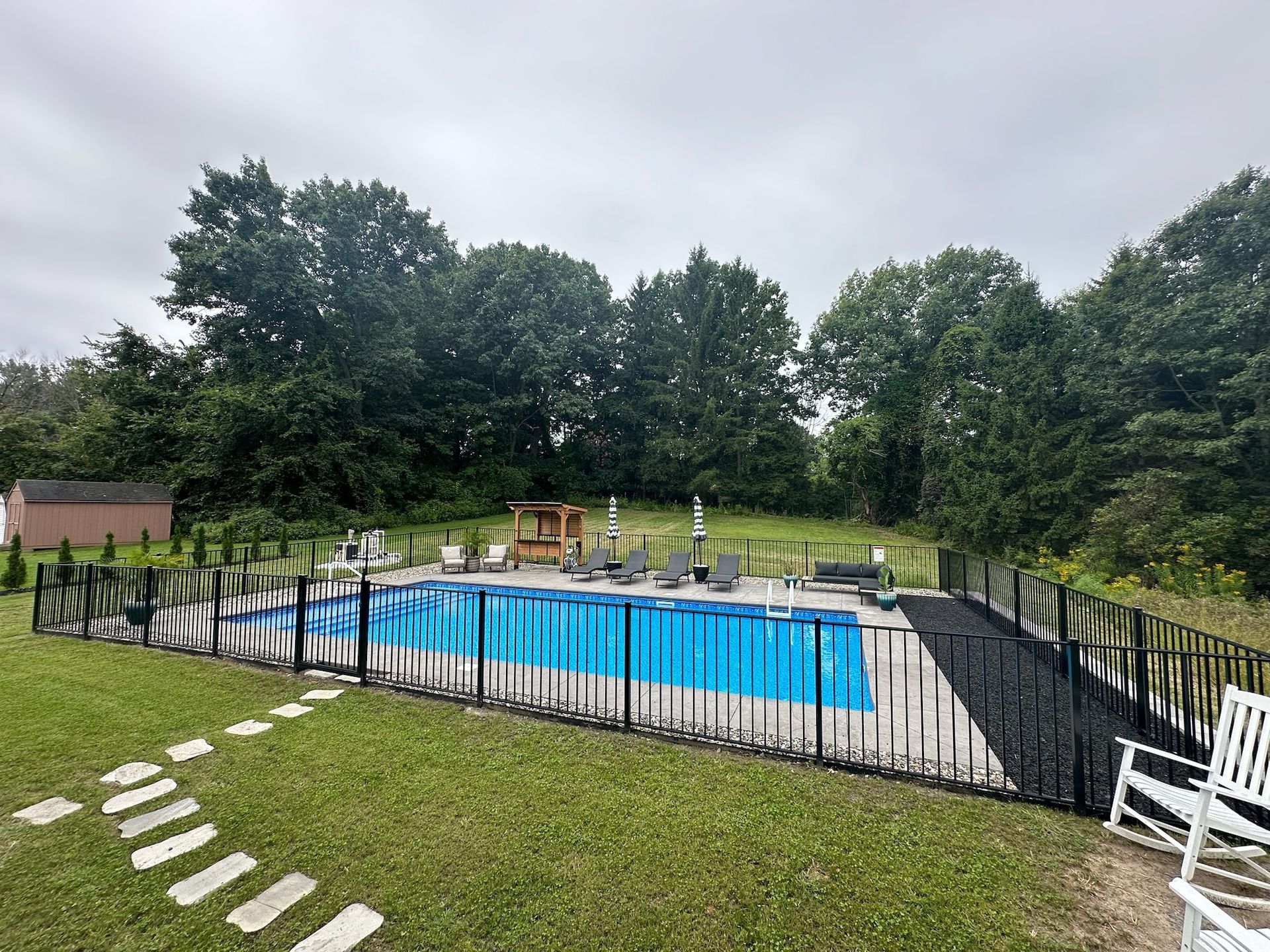 Above-ground pool with blue cover, surrounded by wooden deck and black fence, lush green grass and trees in the background.