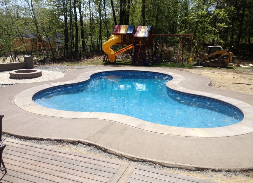 Swimming pool with concrete surround, play structure in the background, and fire pit to the left.