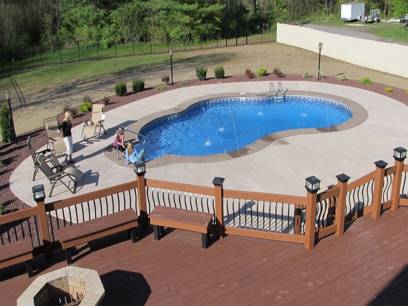 Swimming pool with deck, brown railing, concrete patio, green yard, and two people.