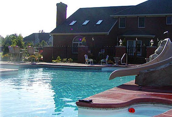 Swimming pool in front of a brick house with a slide and lounge chairs. Blue water with a sunny sky.