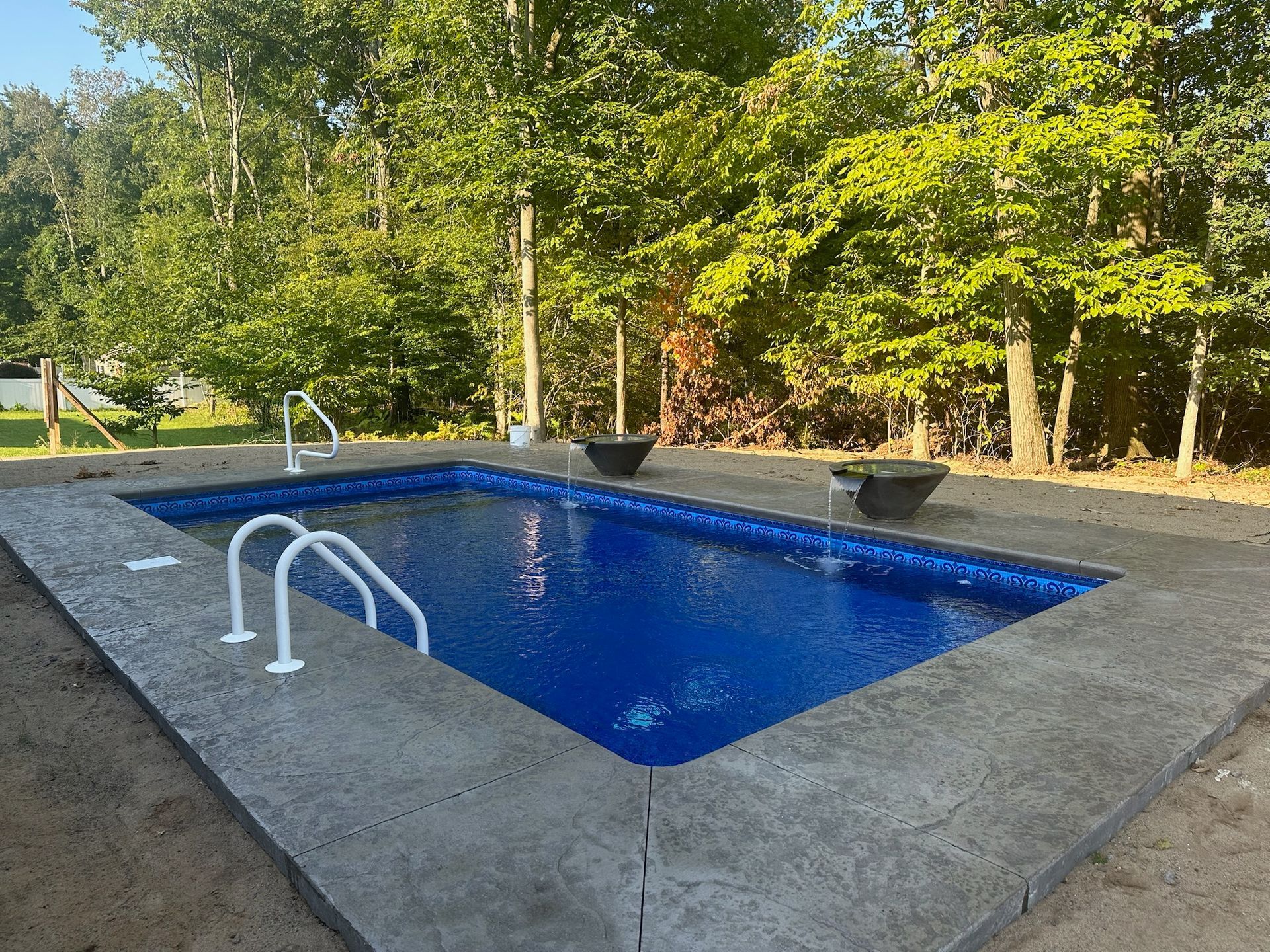 Rectangular in-ground pool with blue water surrounded by concrete patio. Two white ladders. Trees in the background.