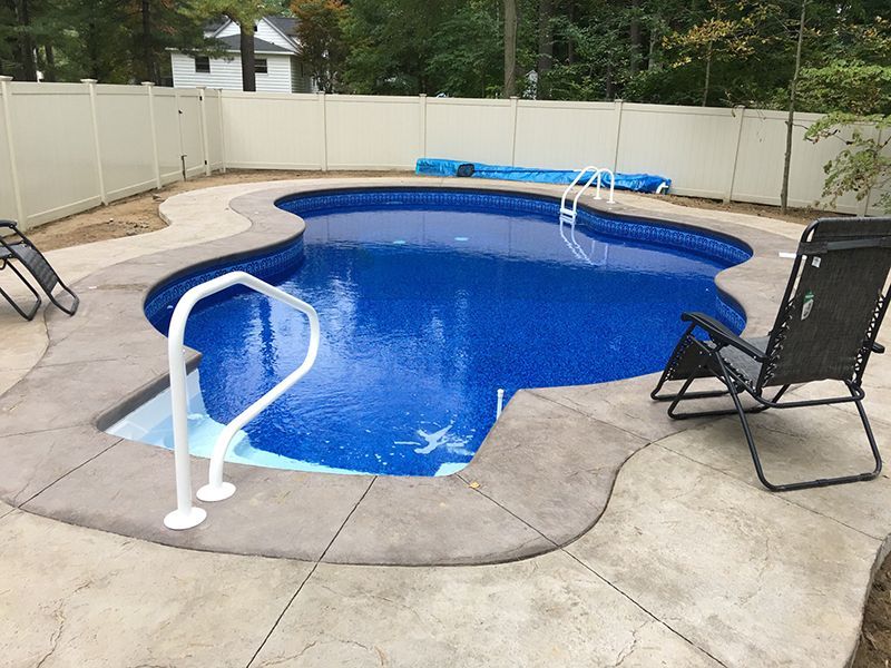 Swimming pool with blue water and concrete deck, enclosed by a white fence. Two chairs sit poolside.
