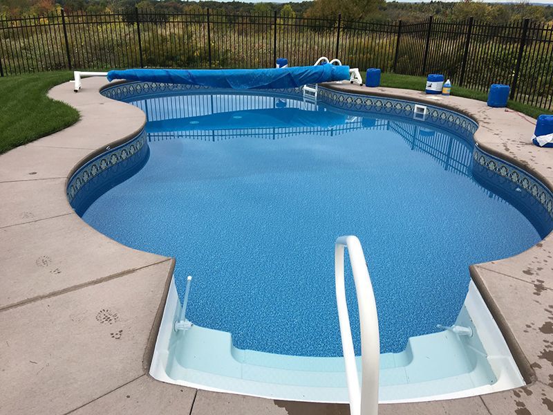 A blue, kidney-shaped swimming pool with a white handrail and a blue cover, set in a grassy backyard.