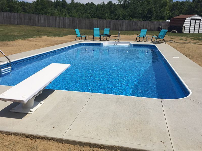 Rectangular swimming pool with diving board, surrounded by concrete. Blue chairs sit nearby on a lawn.