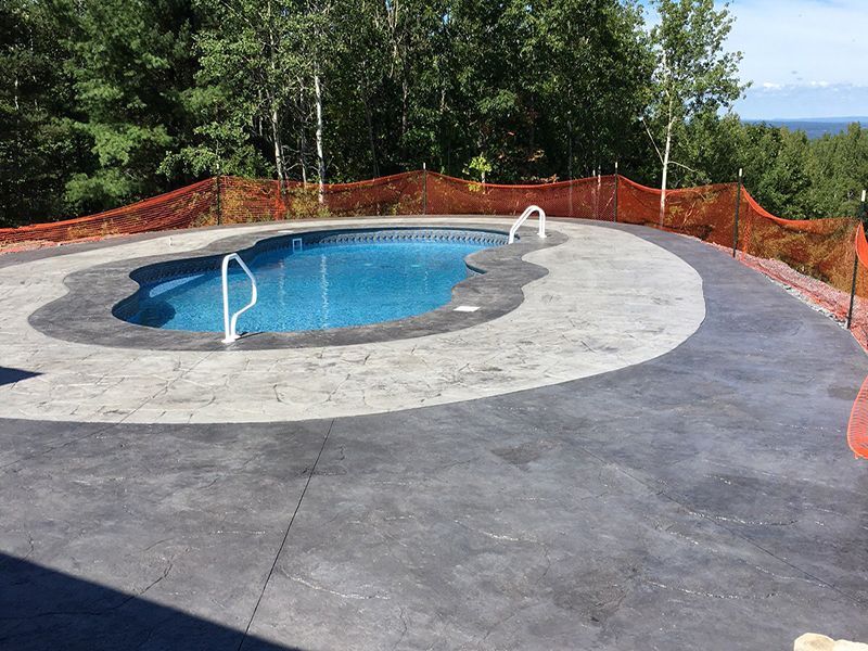 A concrete patio surrounds a blue, kidney-shaped pool. Orange safety fencing lines the edge with trees in the background.