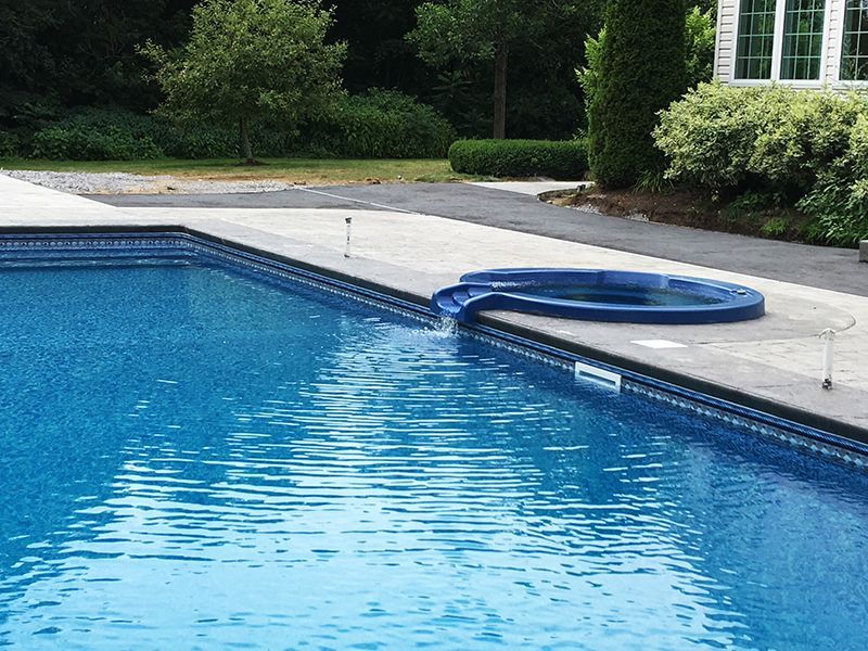 Pool with attached hot tub, blue water, gray concrete surround, green landscaping, sunny day.