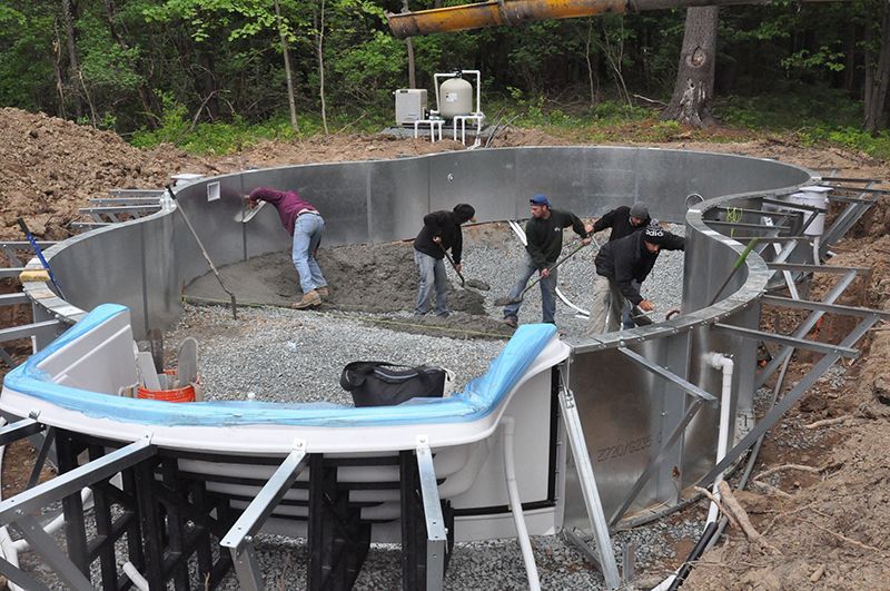 Construction workers installing a pool. They are in the pool's metal shell, shoveling gravel. Trees are in the background.
