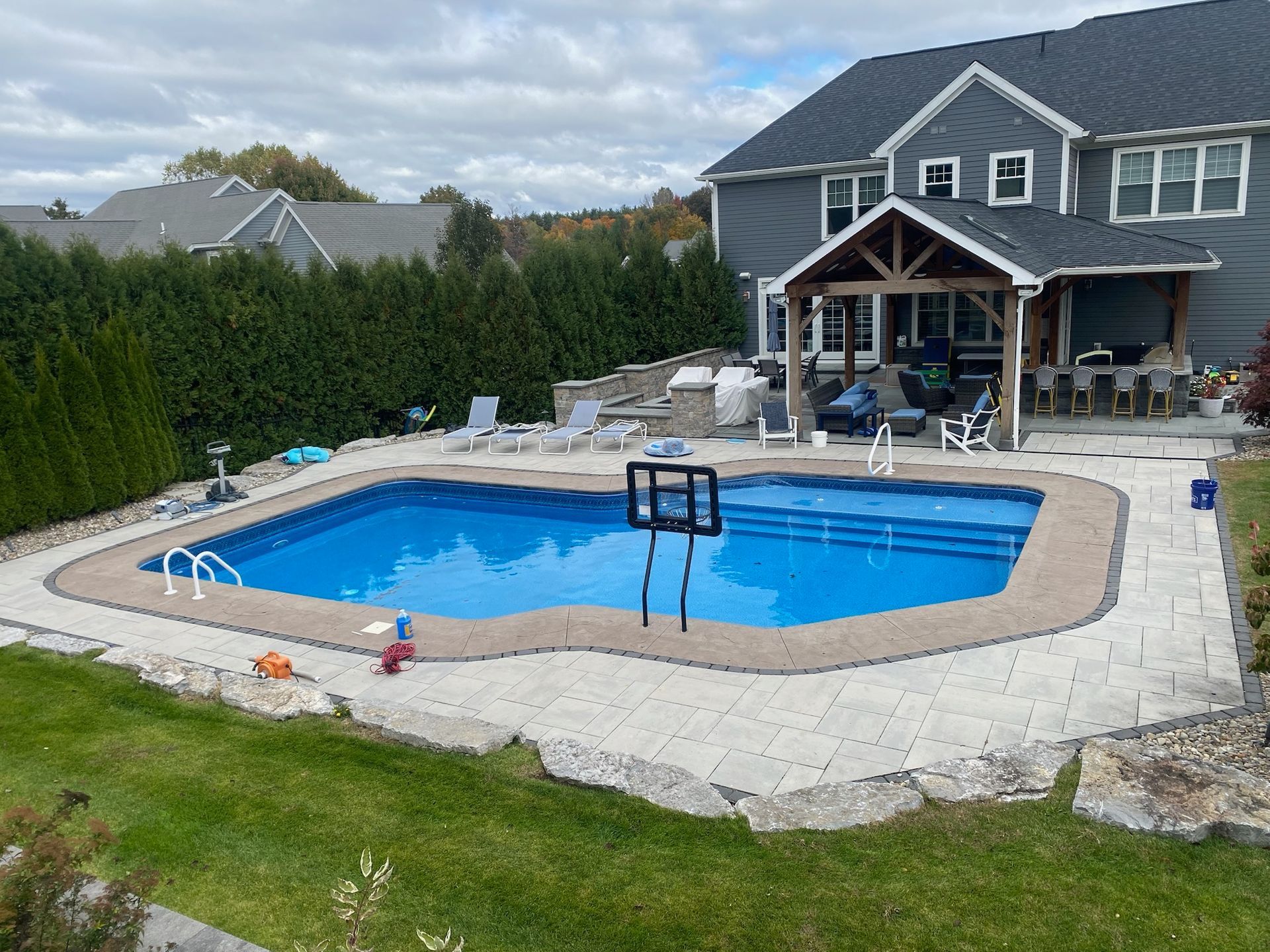 Backyard with a blue pool, stone patio, and a covered seating area near a large house on a cloudy day.