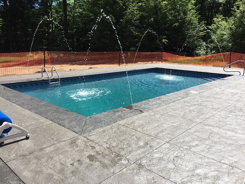 Rectangular pool with water fountains, surrounded by concrete patio and orange safety fence. Trees in the background.