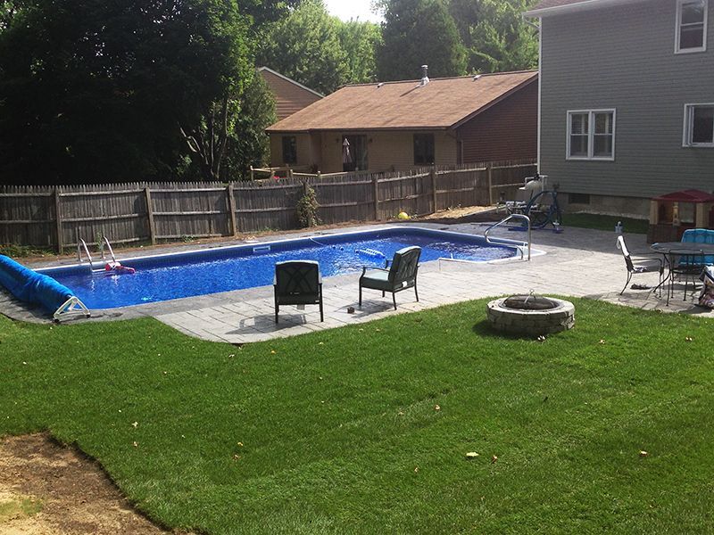 Backyard with rectangular pool, patio, lawn, fence, and houses in the background on a sunny day.