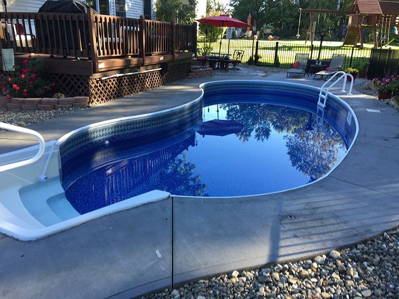 Curved blue swimming pool with concrete surround, deck, and red umbrella in background.