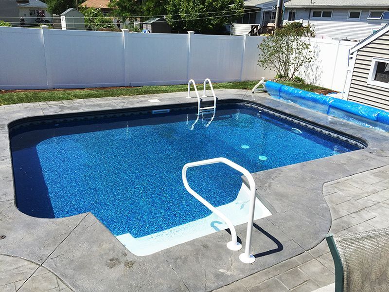 Rectangular in-ground pool with blue water and white ladder. Concrete patio. White fence in the background.