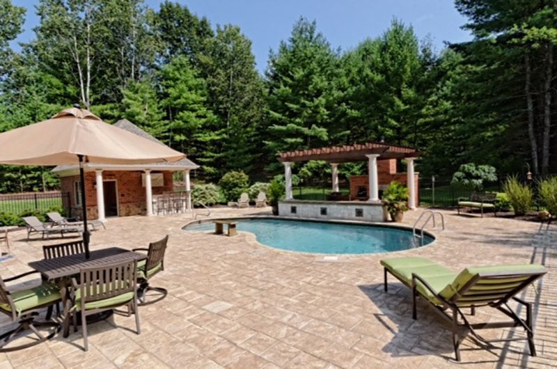 Poolside patio with pool, bar, pergola, lounge chairs, and umbrella-covered table. Surrounded by trees, under a blue sky.