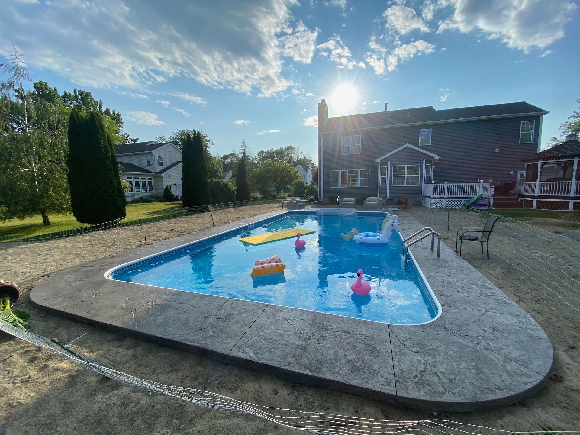 Backyard pool with floats and stamped concrete edging, house in background under a sunny sky.