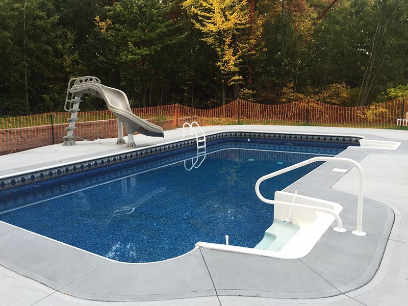 Outdoor swimming pool with a slide and a shallow entry area, surrounded by a concrete deck and a wooden fence.