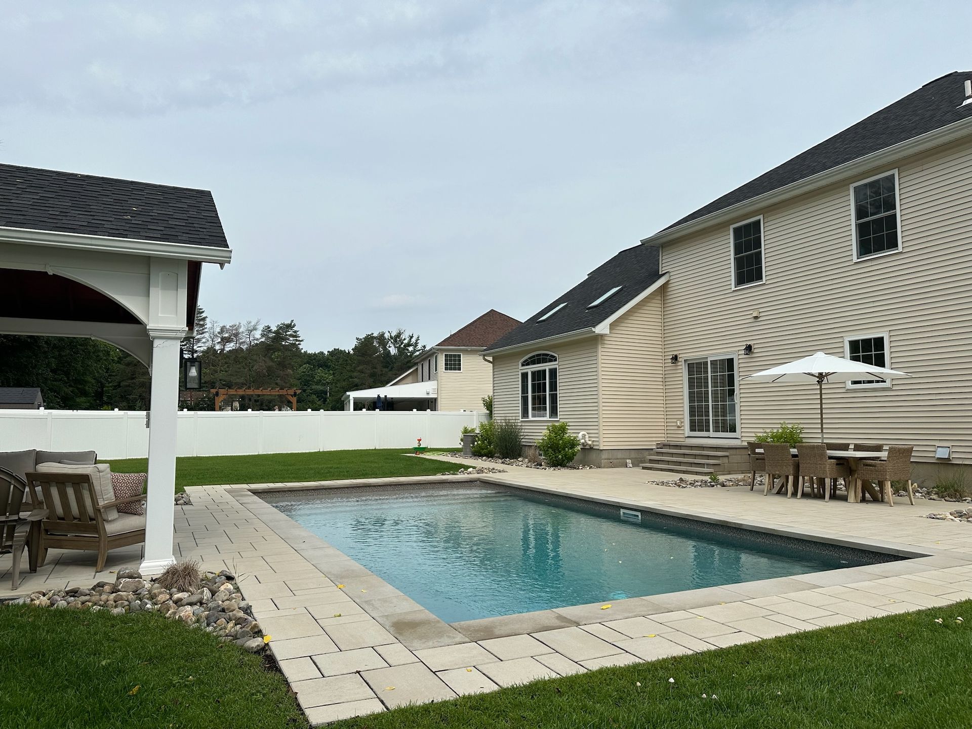 Backyard with a swimming pool, patio, gazebo, and two-story house under a cloudy sky.