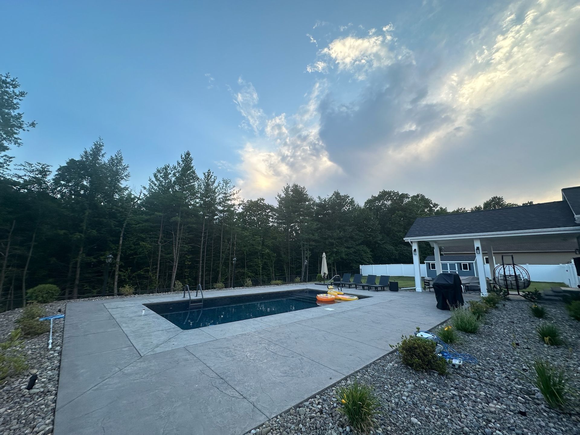 Swimming pool with surrounding patio, trees, and cloudy sky.