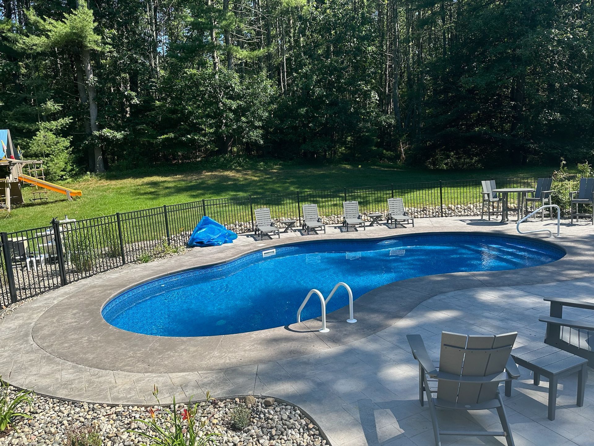Oval-shaped swimming pool surrounded by concrete patio with lounge chairs. Green lawn and trees in the background.