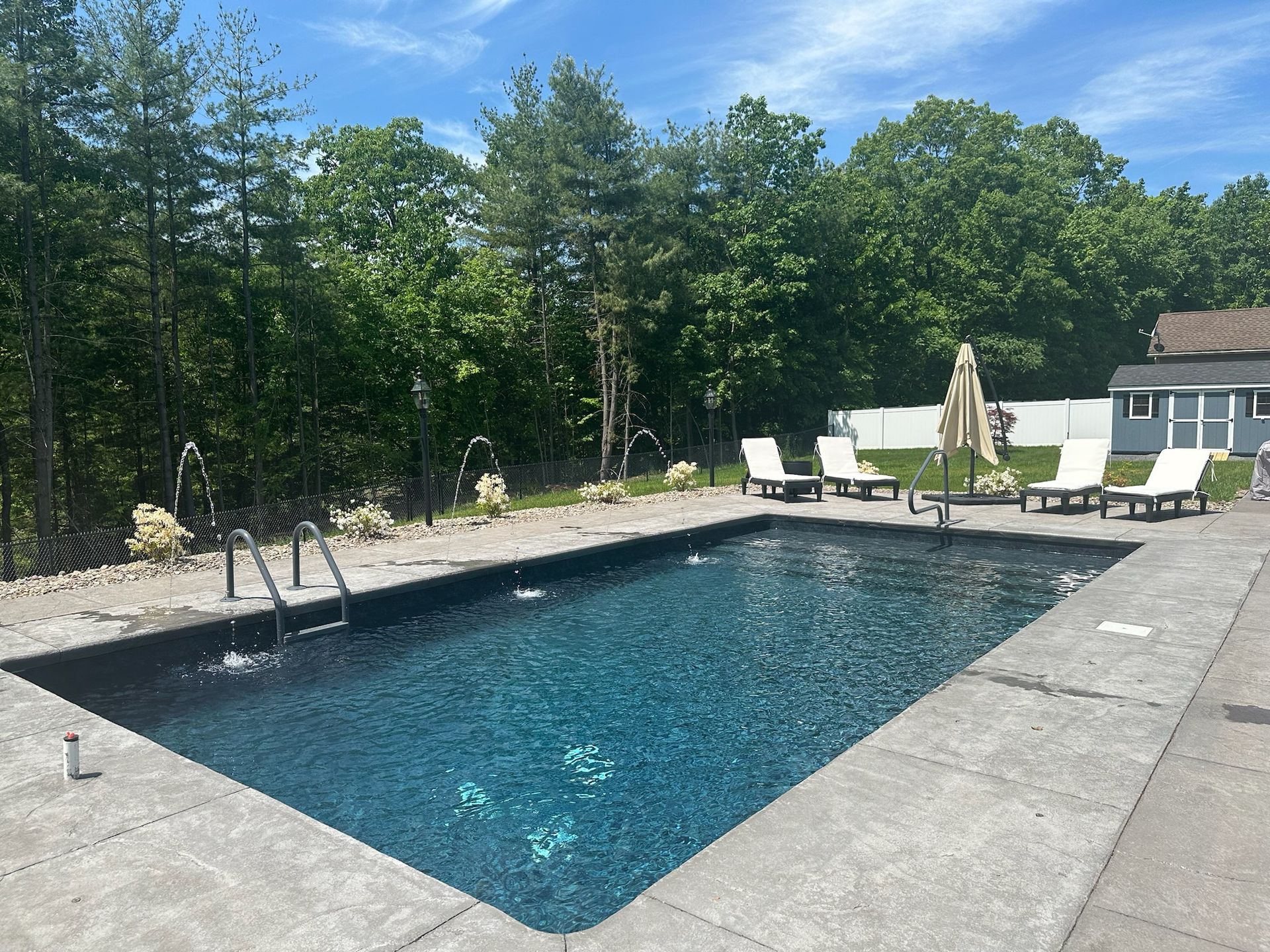 Outdoor pool with fountains, surrounded by concrete and lounge chairs. Trees in the background, blue sky.